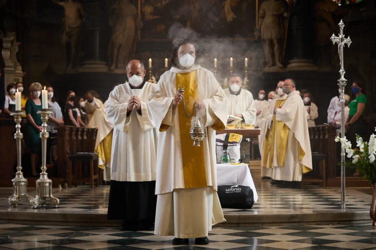 Im warmen Kerzenlicht einer alten Kirche schreitet ein Priester im creme‑goldenen Gewand durch zarten Weihrauch, flankiert von Messdienern und mit Mundschutz maskierten Gläubigen – ein stiller, feierlicher Moment voller Andacht.