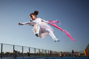 Kind mit rosa Tuch vor blauem Himmel im Sprung auf einer Trampolinanlage
