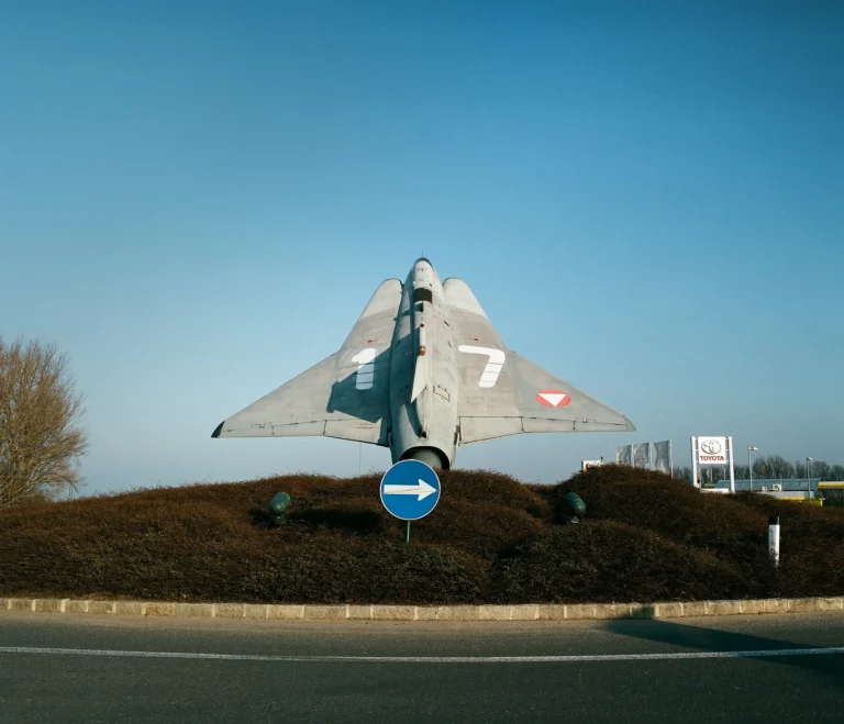 Ein grauer Oldtimer‑Jet streckt sich im goldenen Morgenlicht aus der Mitte eines Kreisverkehrs nach oben, darunter ein blaues Richtungsschild, umgeben von braunen Sträuchern und weitem, wolkenlosem Himmel.