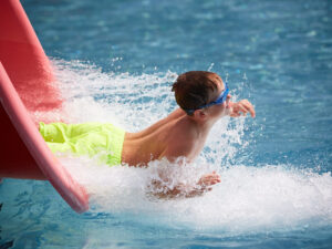 Ein Junge mit Schwimmbrille schießt in einer Gischt kühlen Wassers aus einer Rutsche in ein Schwimmbecken des Wiener Gänselhäufels.
