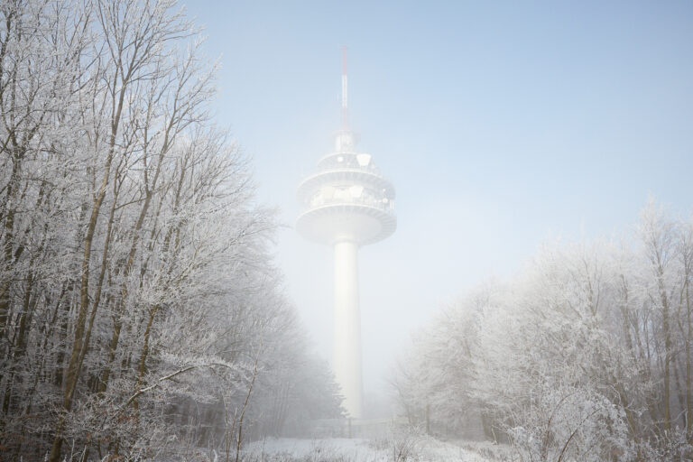 Ein schlanker Fernsehturm taucht aus einem Schleier aus Raureif und Nebel auf, umgeben von frostglitzernden Waldkronen unter zartblauem Winterhimmel.