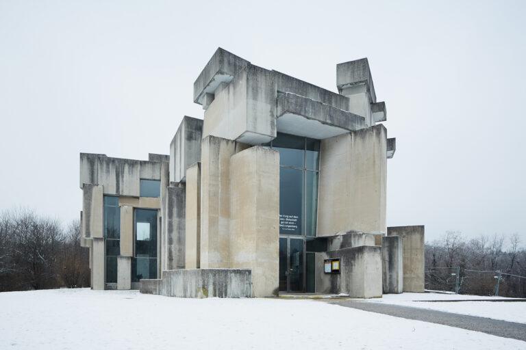 Die kantige Wotruba‑Kirche steht wie eine stille Steinskulptur im leichten Schnee, ihre hellen Betonquader ragen ruhig in den blassgrauen Winterhimmel.